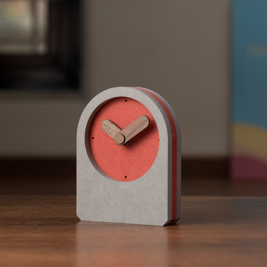 A square-shaped, red and gray wooden table clock with visible gears and a cuckoo bird design on the hour hand, standing on a flat surface.
