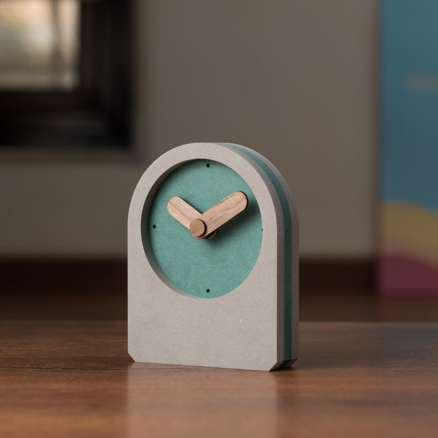 A square-shaped table clock with oval wooden hands, predominantly in green color, with a white background and a black minute hand.