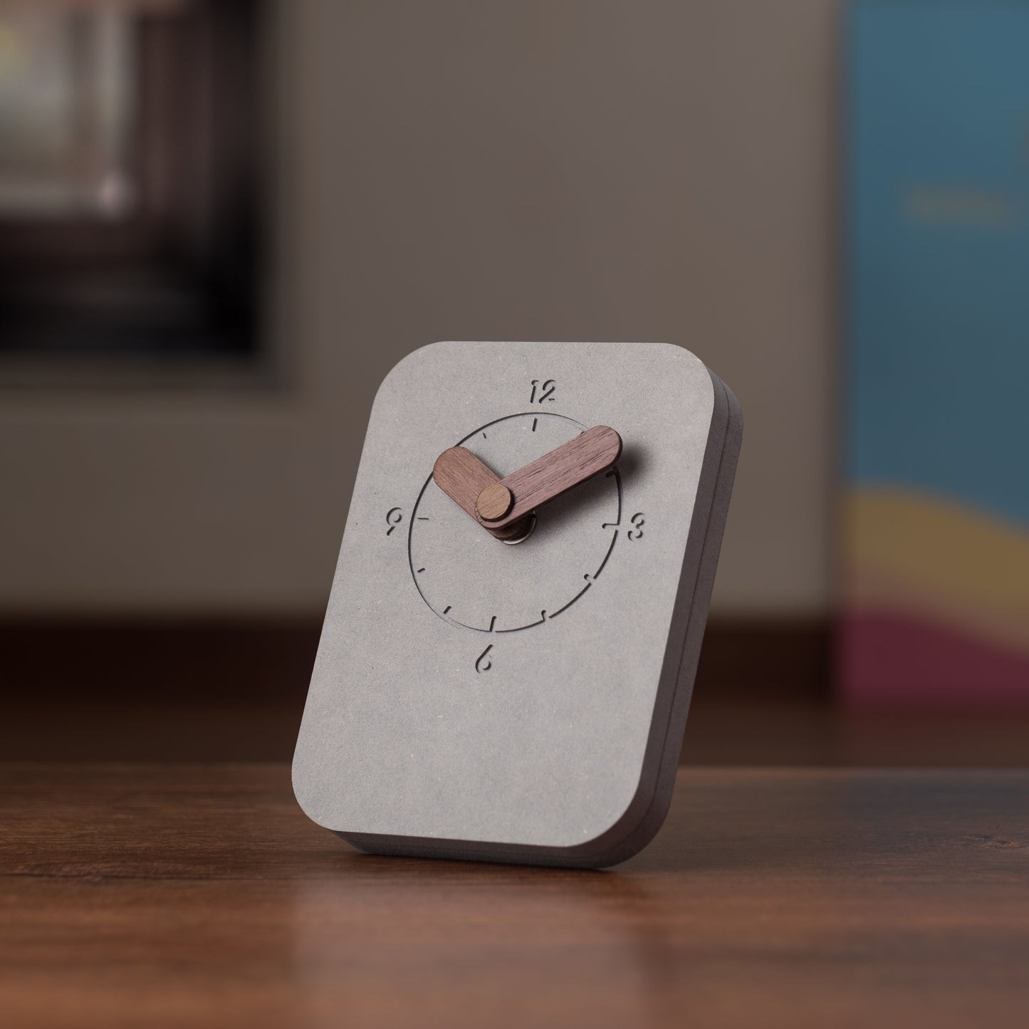 A rectangular grey table clock with wooden hands, displaying time with black numerals, placed on a wooden surface.